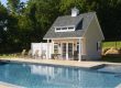 Modern prefab pool house beside a luxury backyard swimming pool with outdoor lounge furniture and landscaped garden.