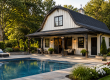 Luxury barn-style pool house with a covered porch beside a modern backyard swimming pool.